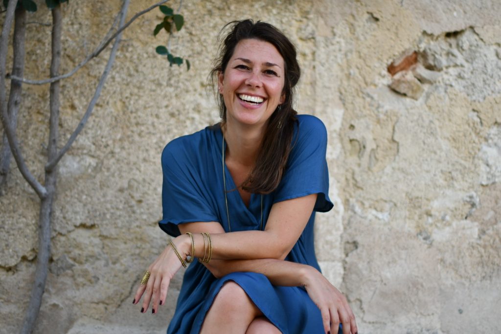 Portrait photo of Kanika Frings - Founder of Dima Mallorca Mystery School smiling in a blue dress in front of Dima wall